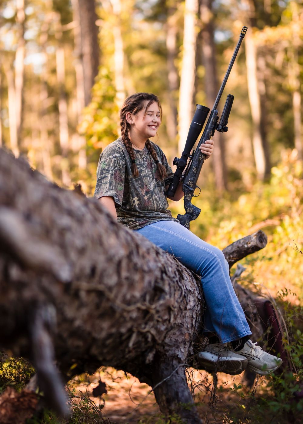 Hunter in camouflage clothing sits on fallen log with rifle during autumn hunt in sunlit forest.