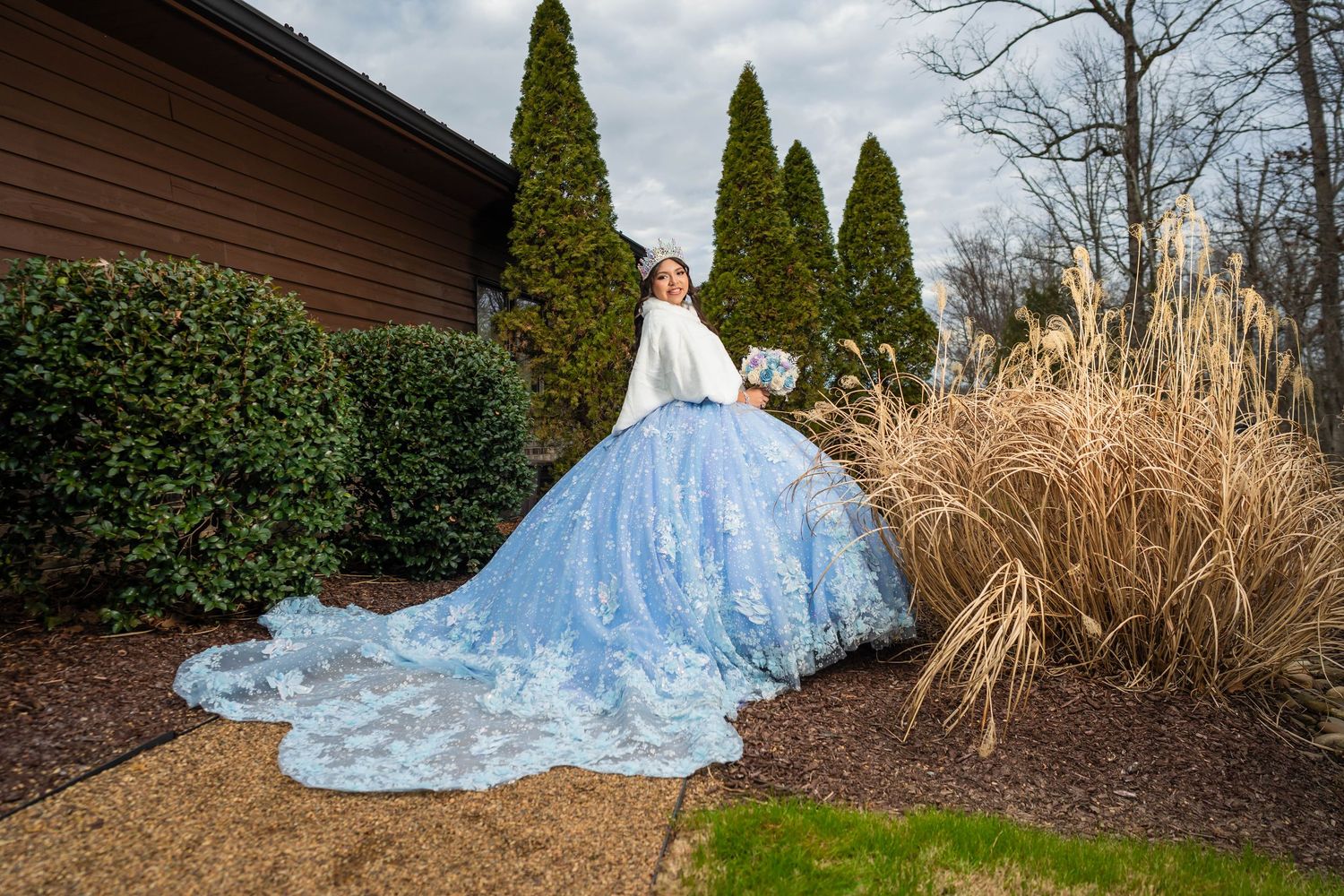 A quinceañera in a flowing light blue wedding gown poses in a garden setting with evergreen trees and ornamental grasses.