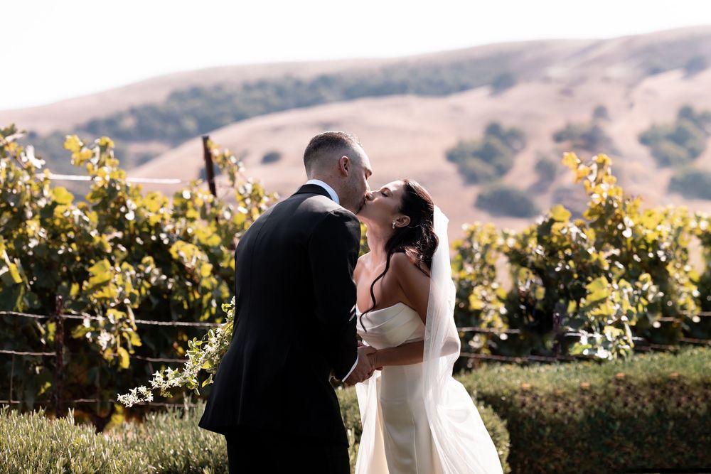 A couple shares a romantic embrace among sun-drenched vineyard rows during a wedding photoshoot at sunset.