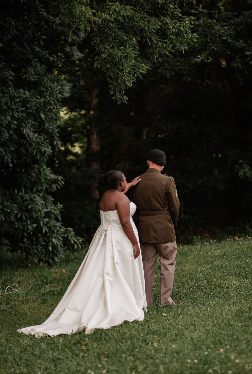 Wedding couple walks together on green lawn with trees in background.