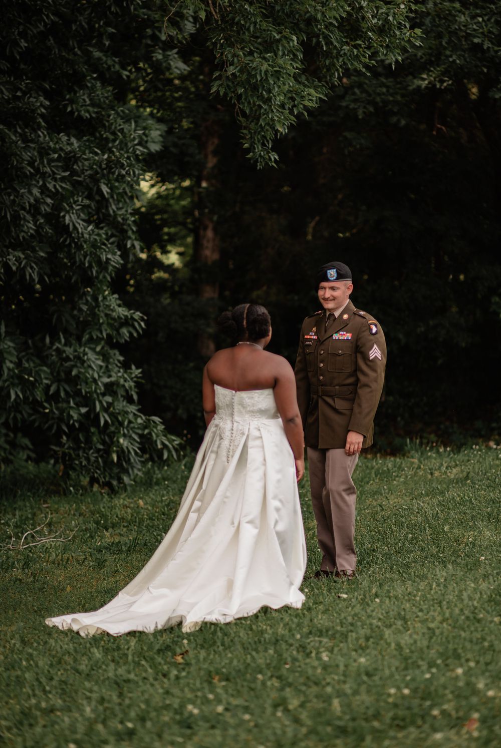 A couple stands together outdoors, with the bride's elegant white gown flowing across the grass.
