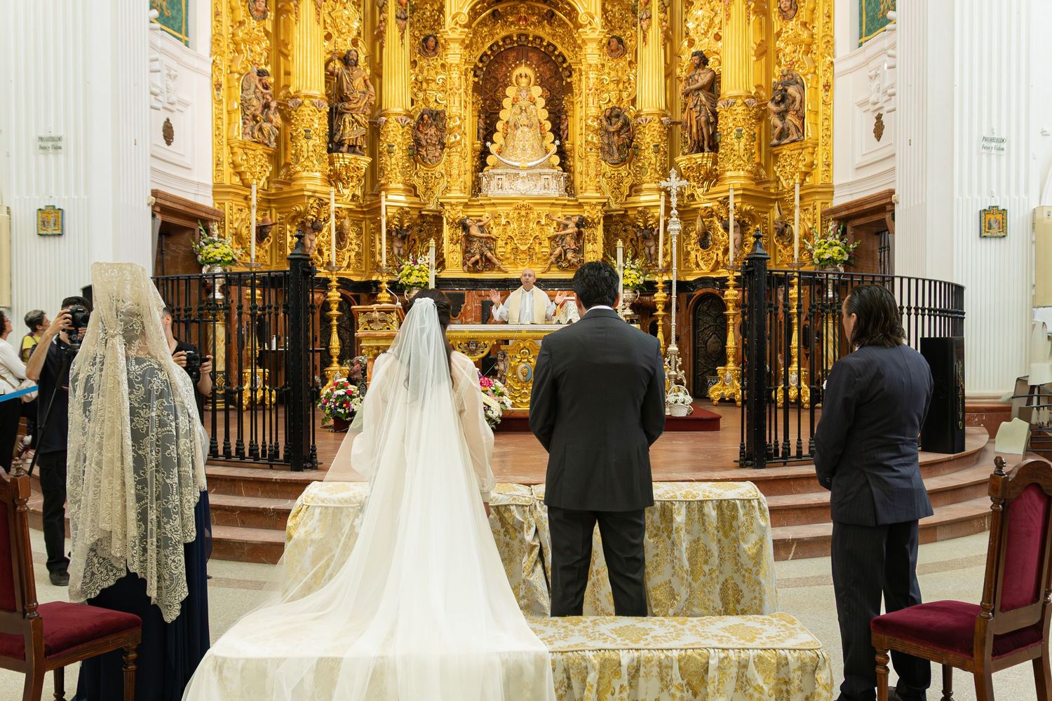 Wedding ceremony in an ornate golden baroque church interior with long-trained white wedding dress and guests in attendance.