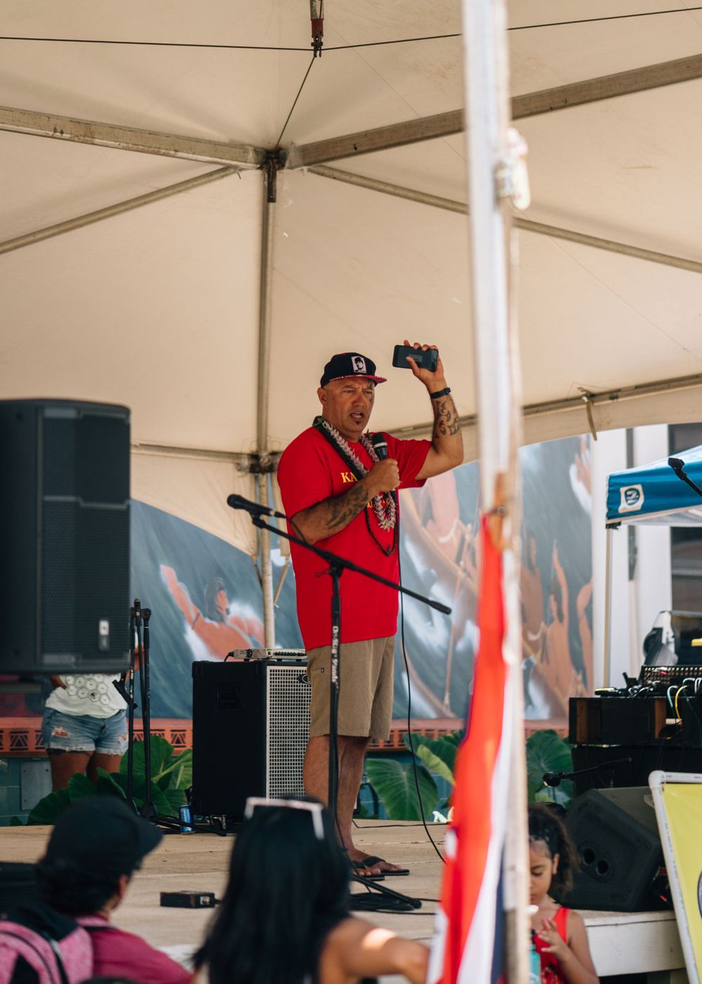 A performer in a red shirt and baseball cap speaks into a microphone under a white tent at an outdoor event.