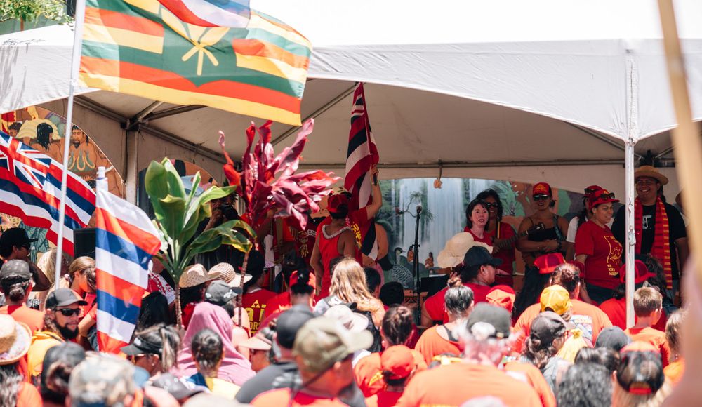 Hawaiian flags wave above a crowded outdoor celebration with people in red attire.