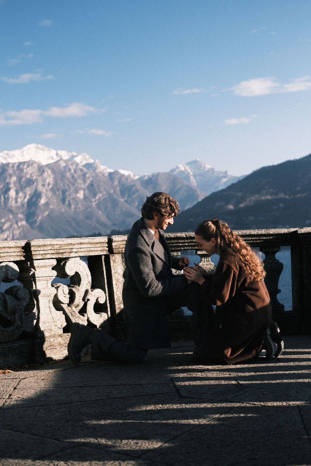Two people share an intimate moment on a stone balcony overlooking majestic snow-capped mountains on a sunny day.