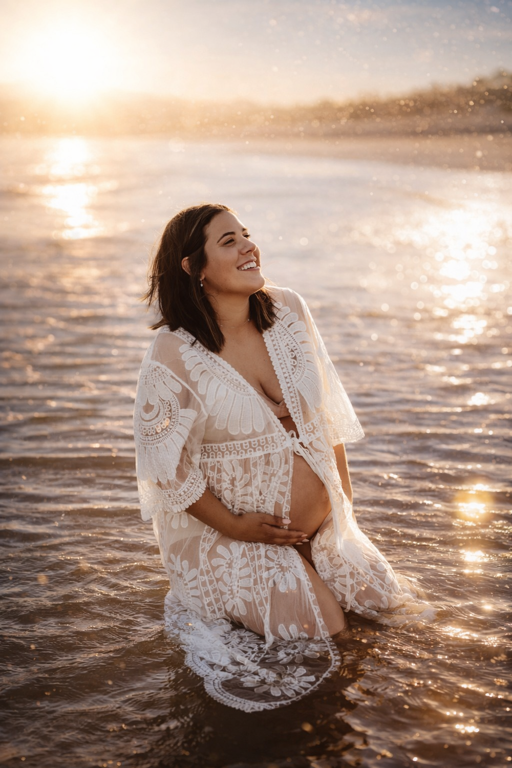 Woman in white lace dress laughing in ocean water during golden sunset.