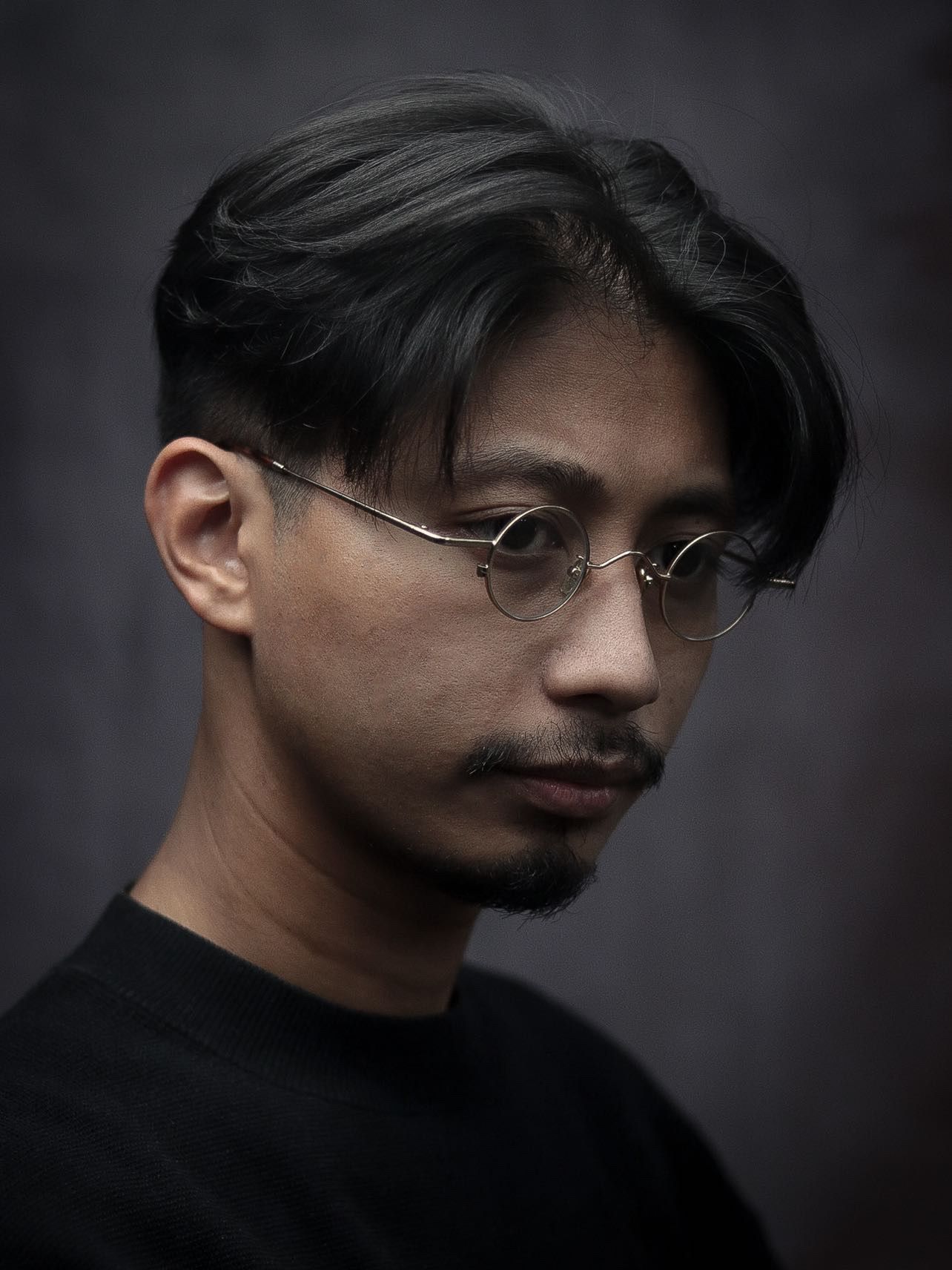 Moody portrait of a person in black wearing round glasses against a dark background.