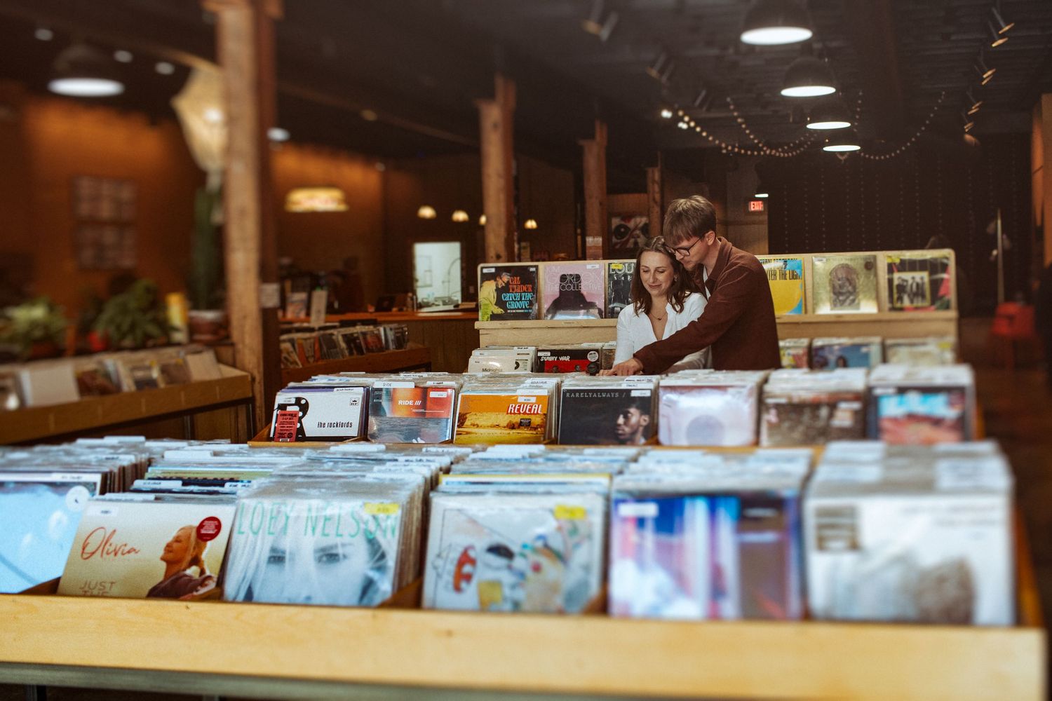 A couple browse through vinyl records in a well-lit record store with wooden shelving and display cases. Indianapolis, Indiana engagement