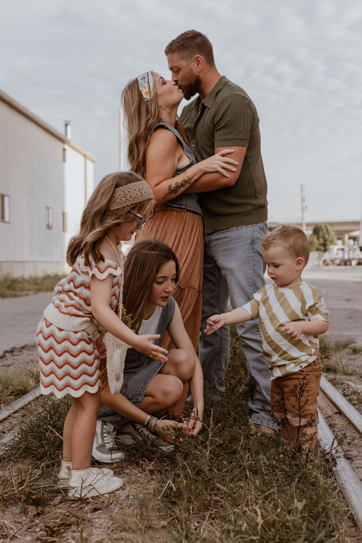A Kansas City family photo session featuring a couple sharing a kiss while their three children play on the ground in front of them.