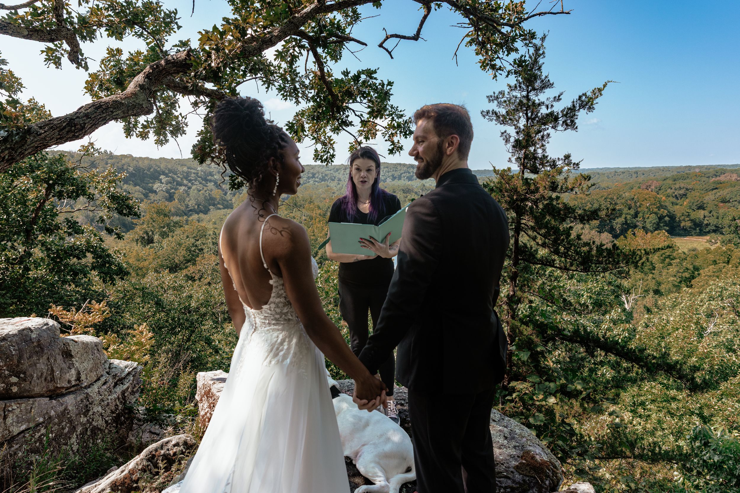 Couple exchanges vows outdoors on rocky overlook with lush green valley backdrop, officiant holding ceremony book.