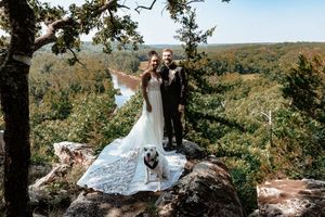 Bride and groom stand on rocky overlook with their white dog, surrounded by lush forest and scenic valley views.