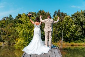 Bride and groom celebrate on a wooden dock overlooking a serene lake surrounded by lush green trees on a sunny day.