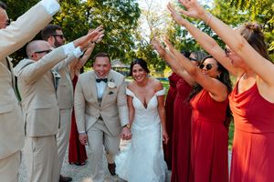 Bride and groom walk through an arch of raised hands formed by wedding party in red dresses and tan suits outdoors.