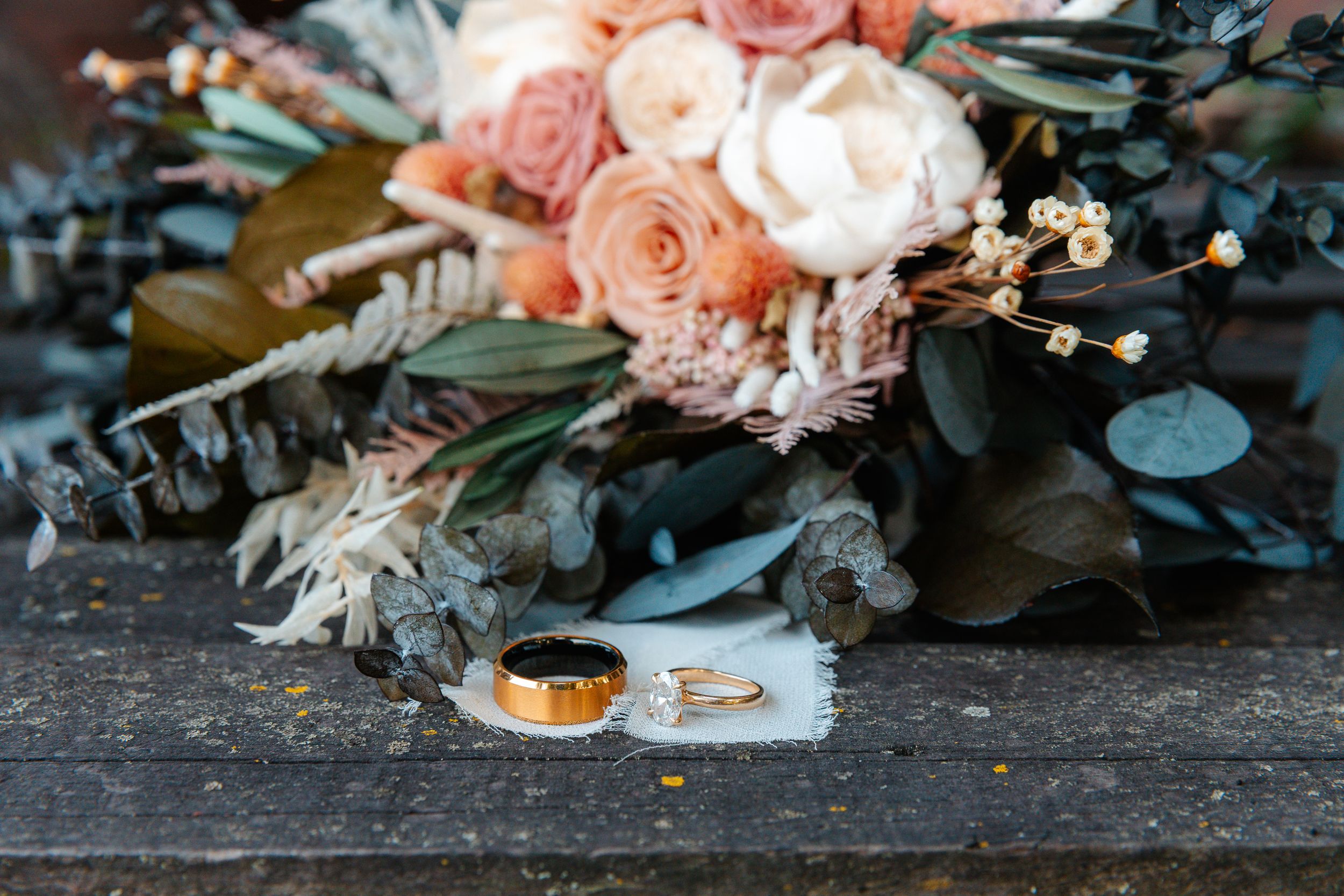 Wedding rings and gold band displayed on lace beside a romantic bouquet of blush pink roses and eucalyptus.