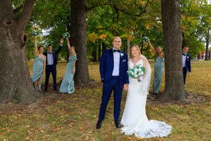 Bride and groom pose before large trees with bridesmaids in sage green dresses and groomsmen celebrating behind them.
