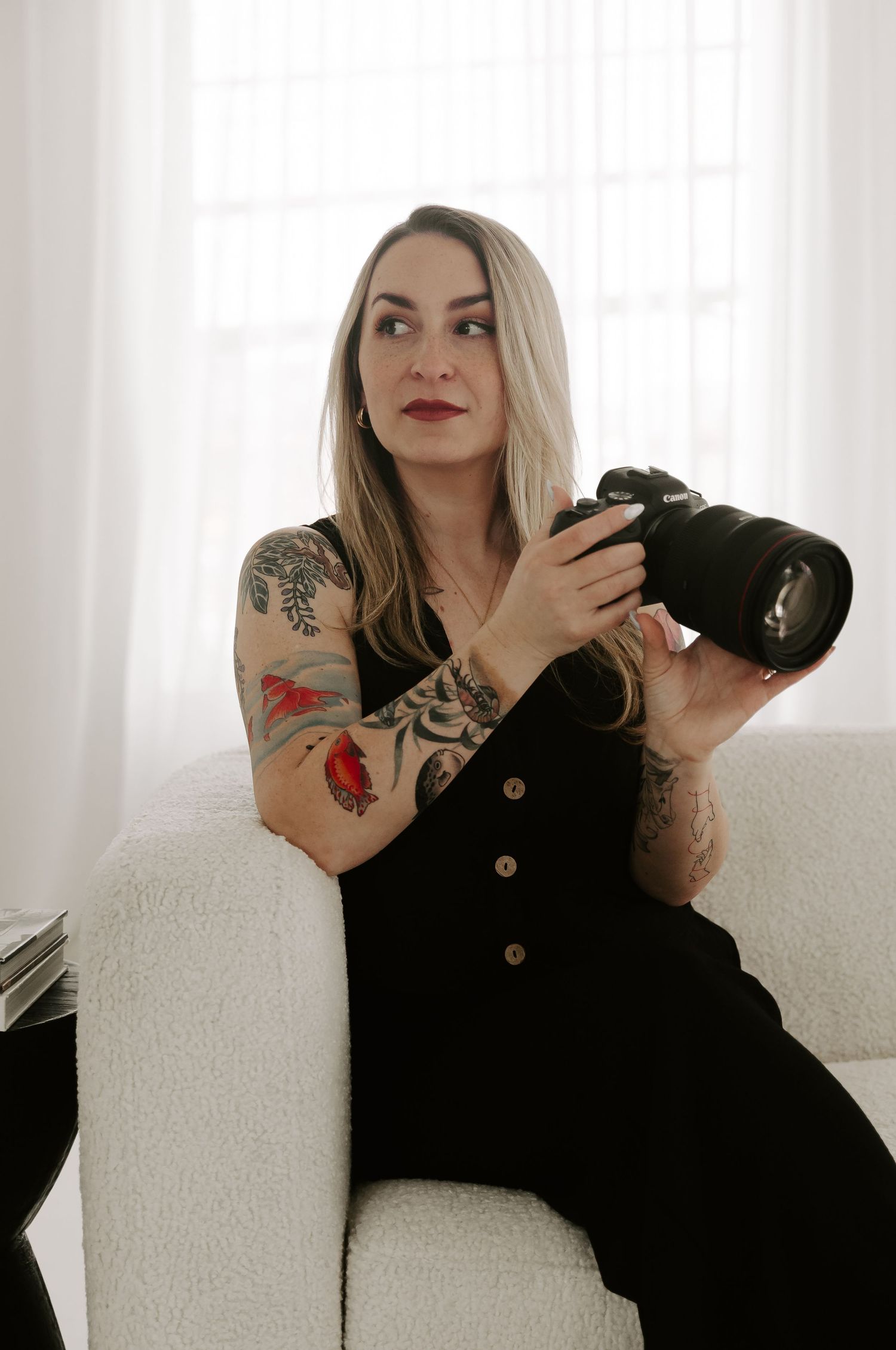 Professional photographer in black dress with colorful arm tattoos holds camera while sitting on white couch.