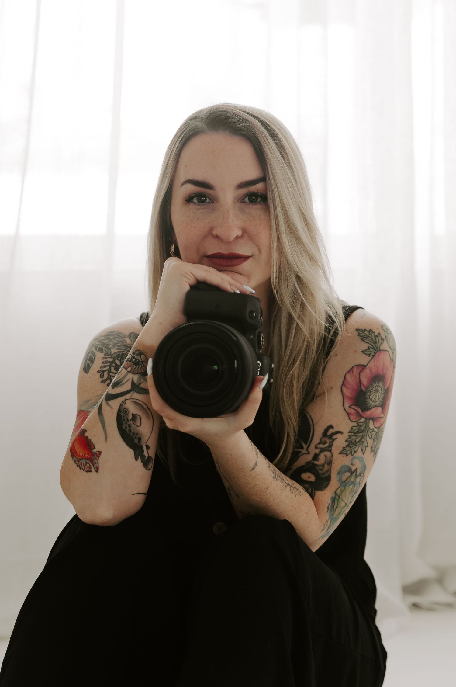 Photographer with tattoos holds professional camera in bright white studio setting.