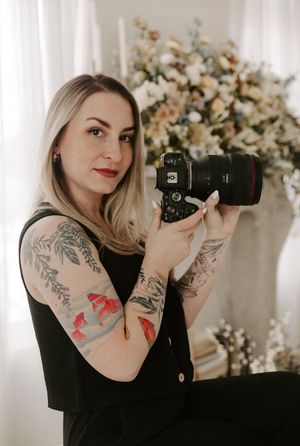 Professional photographer with colorful arm tattoos poses with camera in front of floral backdrop in studio setting.