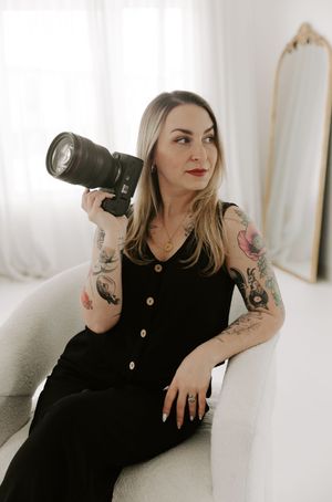 Professional photographer in black dress with tattoos holds camera while posing on white couch in bright studio setting.