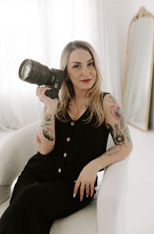 Professional photographer in black dress poses with camera on white couch showing artistic tattoos in lifestyle shoot.