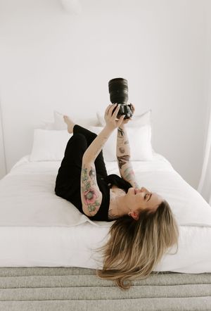 A person with tattoos lies on white bedding while holding a black camera to take photos in a minimalist setting.