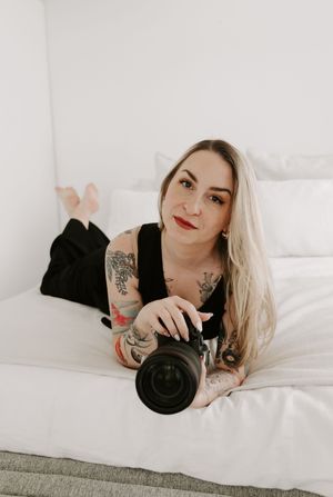 Tattooed photographer lying on white bedding holds professional camera while showcasing creative portrait angles.