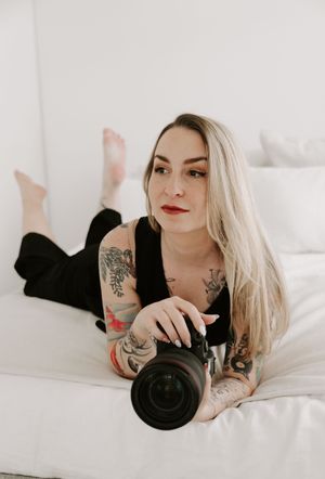 A photographer with blonde hair and tattoos lies on white bedding while holding a camera in three similar poses.