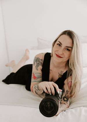 A person in black clothing lying on white bedding with visible tattoos holds a camera lens in a creative pose.