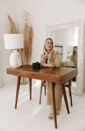 Professional photographer sits at wooden desk with camera and white lamp in minimalist bright studio setting.
