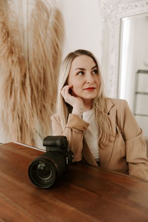 Professional photographer with camera poses playfully in a beige blazer against a warm wooden table setting.