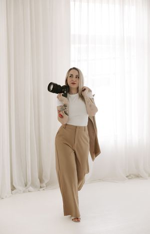 Photographer in tan suit and white top holds professional camera while standing against white curtain background.
