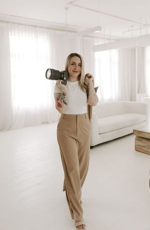 Professional photographer in white top and tan pants holding camera in bright modern studio space.