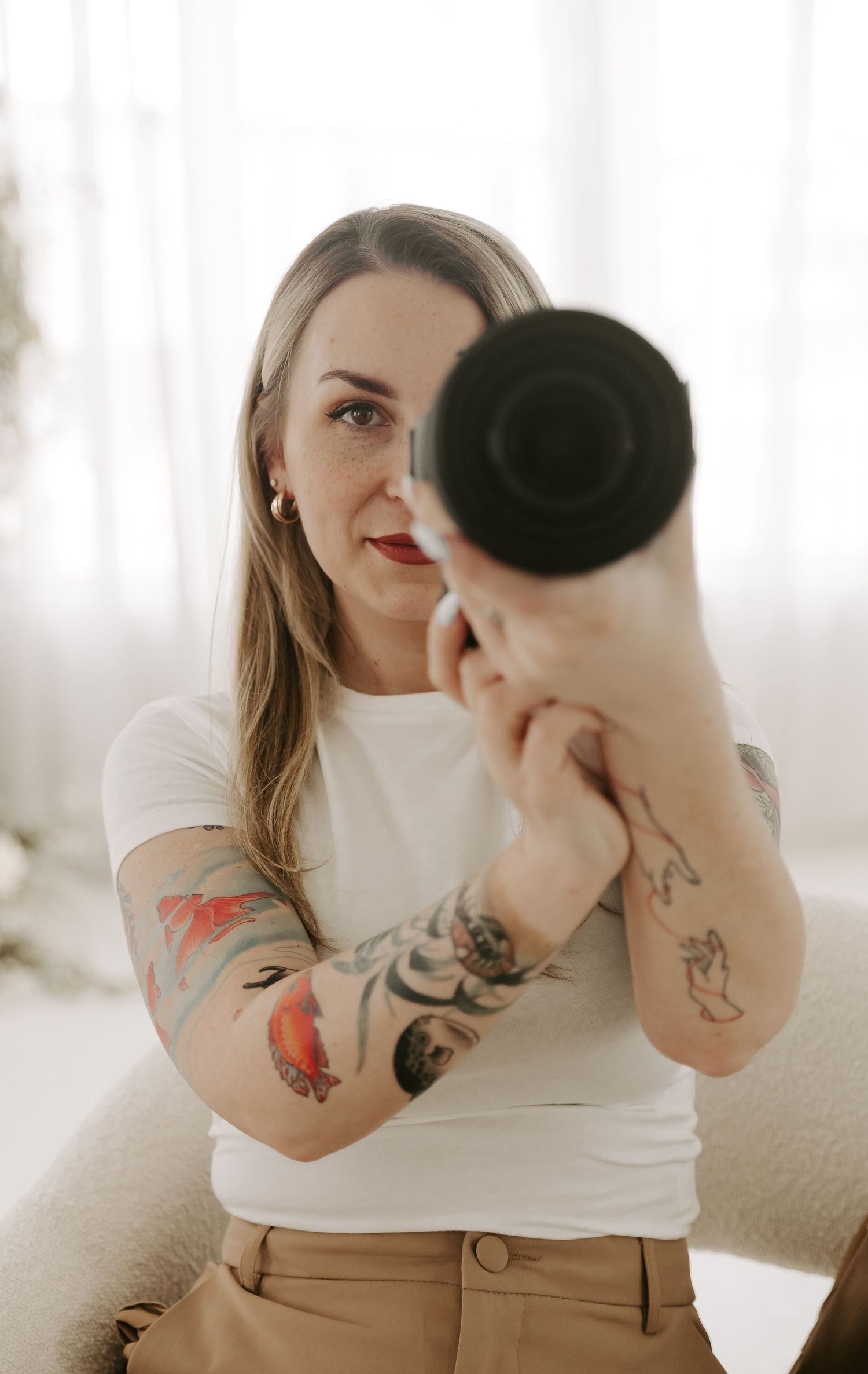 A photographer in a white top with colorful arm tattoos holds up a camera lens while taking a self portrait.