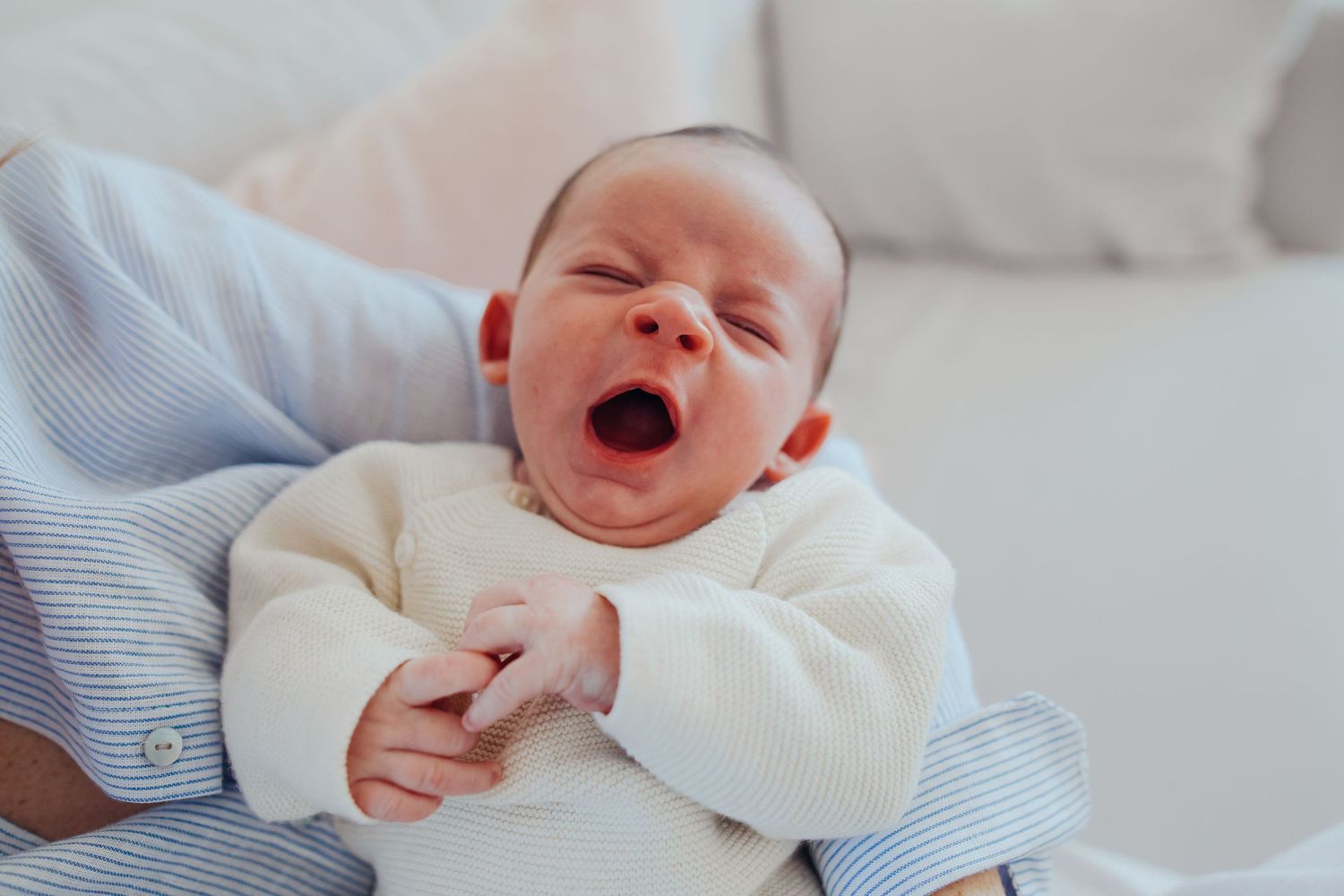 Adorable newborn baby yawning widely while being held in adult arms wearing a white outfit.