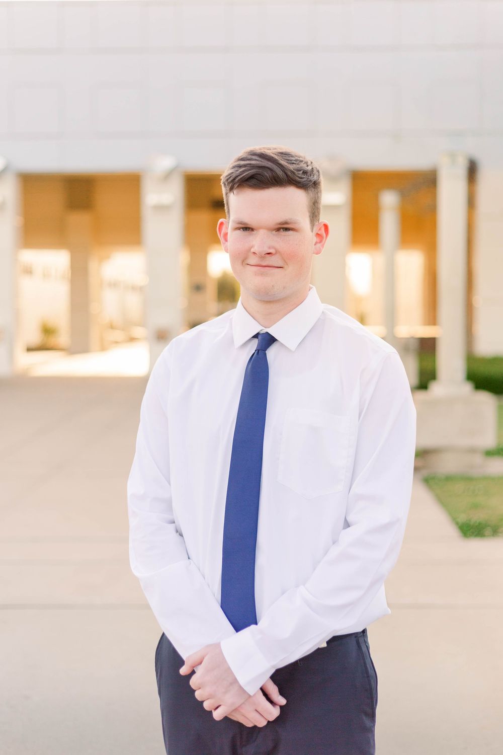 A person wearing a white dress shirt and blue tie stands in front of a light-colored building.