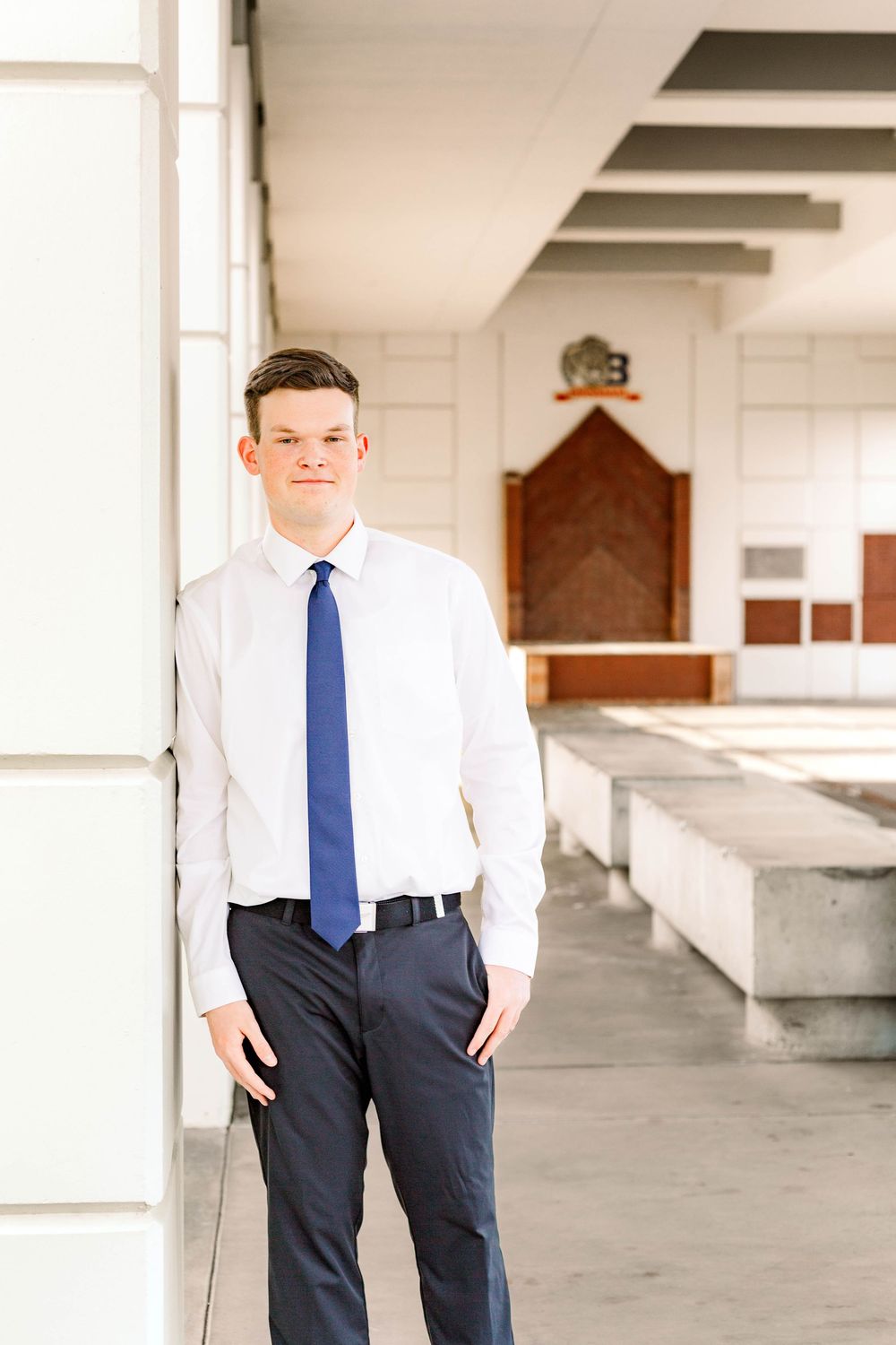 Young man in formal attire poses confidently in white interior space with pillars.