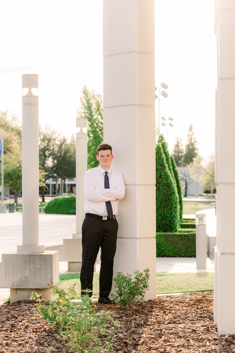 Person in formal attire stands between white columns in an outdoor courtyard with landscaping and mulch.
