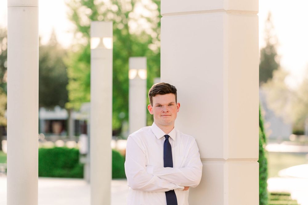 Young person in white dress shirt and navy tie poses confidently with arms crossed against white column outdoors.