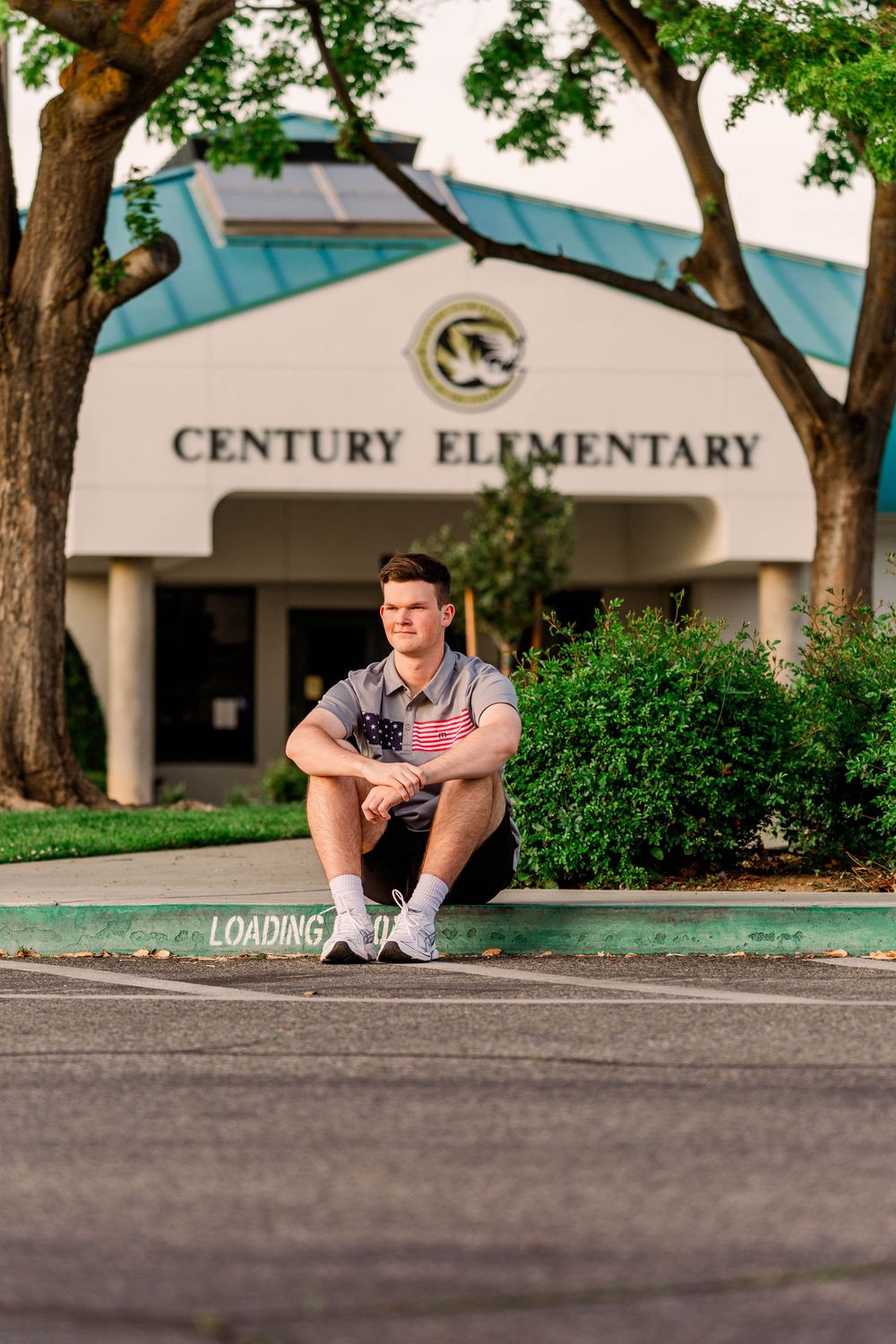 Person sits on pavement outside Century Elementary with trees and bushes in background.