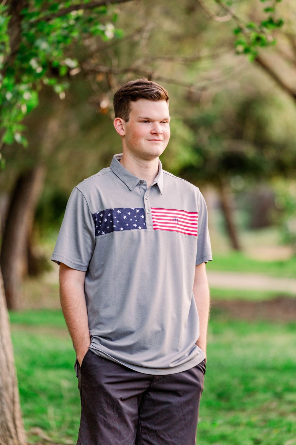 A person in a grey t-shirt with American flag design poses outdoors among green trees.