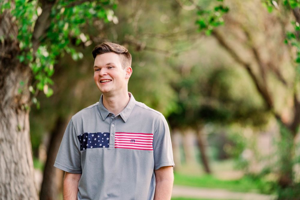 A person wearing a grey American flag t-shirt stands in a park with green foliage in the background.