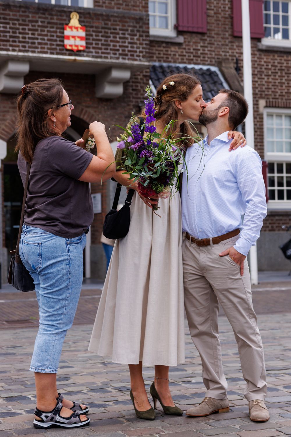 Bruidspaar kust elkaar voor het stadhuis waarvan de moeder van de bruid een foto maakt met haar telefoon - gemeentehuis Boxtel - MaxFotos