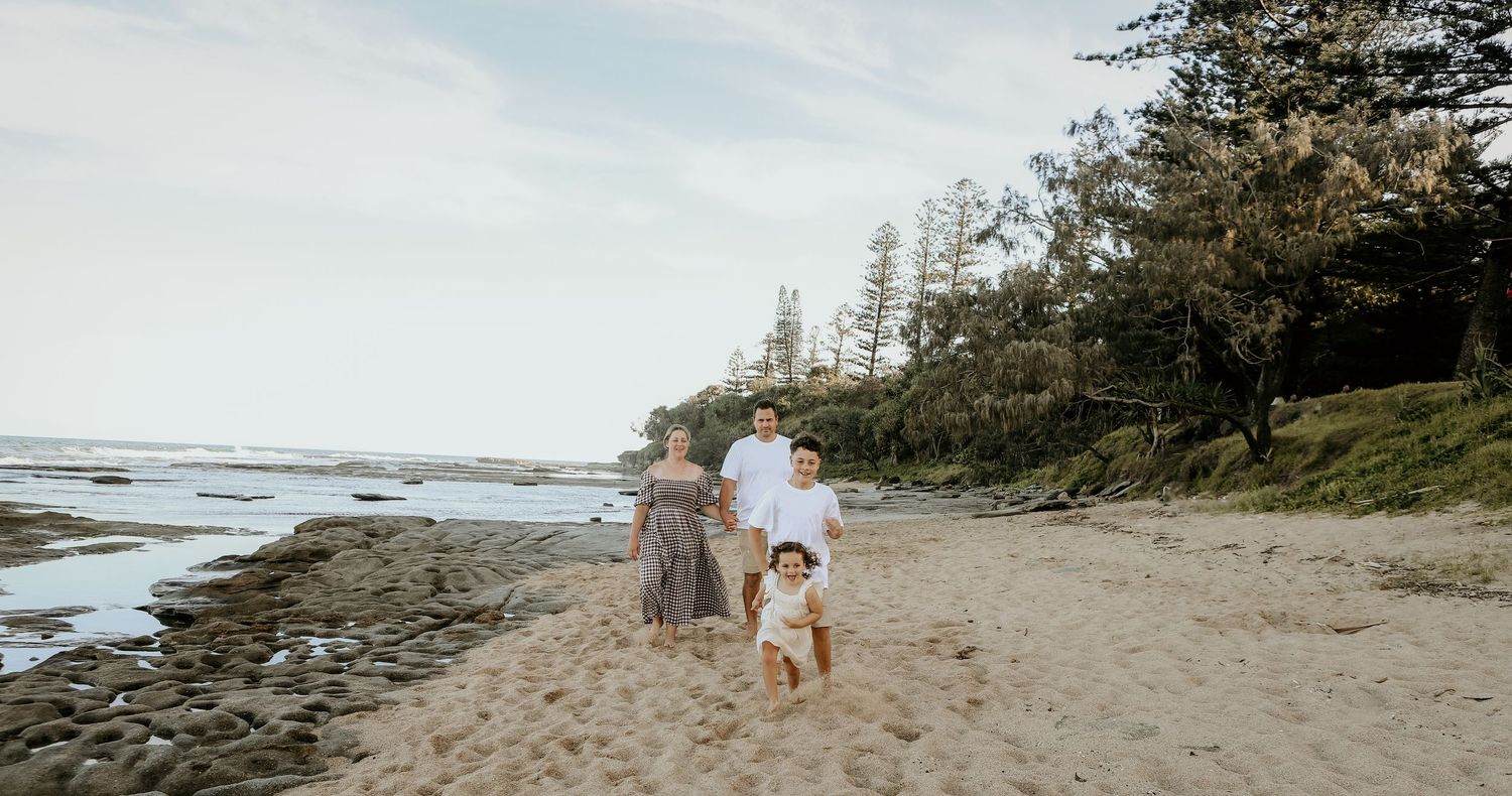 A group walks along a scenic coastal beach with evergreen trees lining the rocky shoreline at sunset.