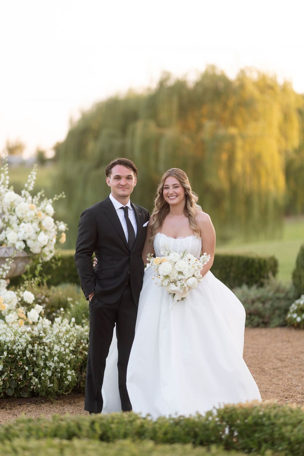 Elegant wedding couple poses in front of lush garden setting at sunset.