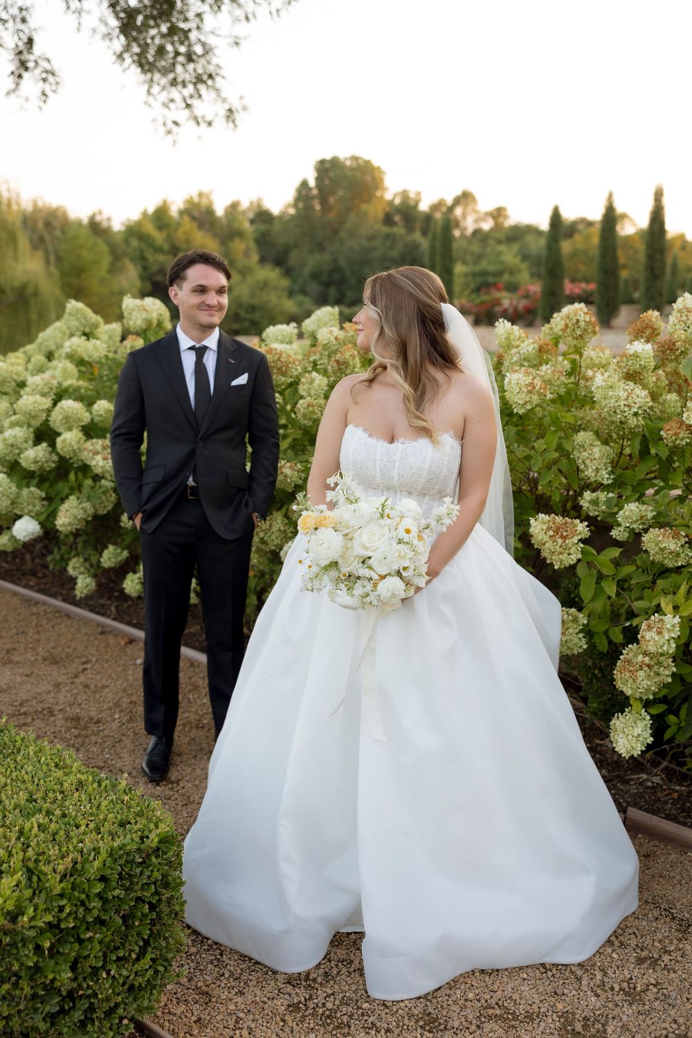 Wedding couple strolls through blooming hydrangea garden during sunset photoshoot.