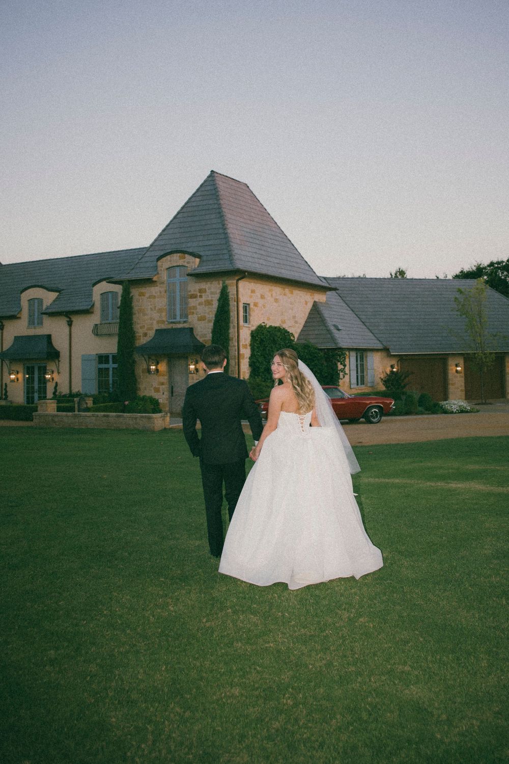 A couple walks together on a green lawn outside a stone building with a white wedding dress flowing in the breeze.