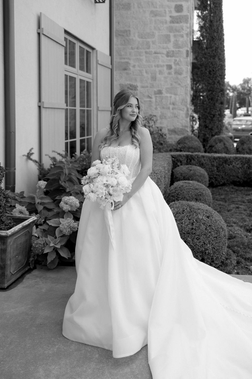 A bride in a flowing white gown poses elegantly on a stone porch with manicured hedges in the background.