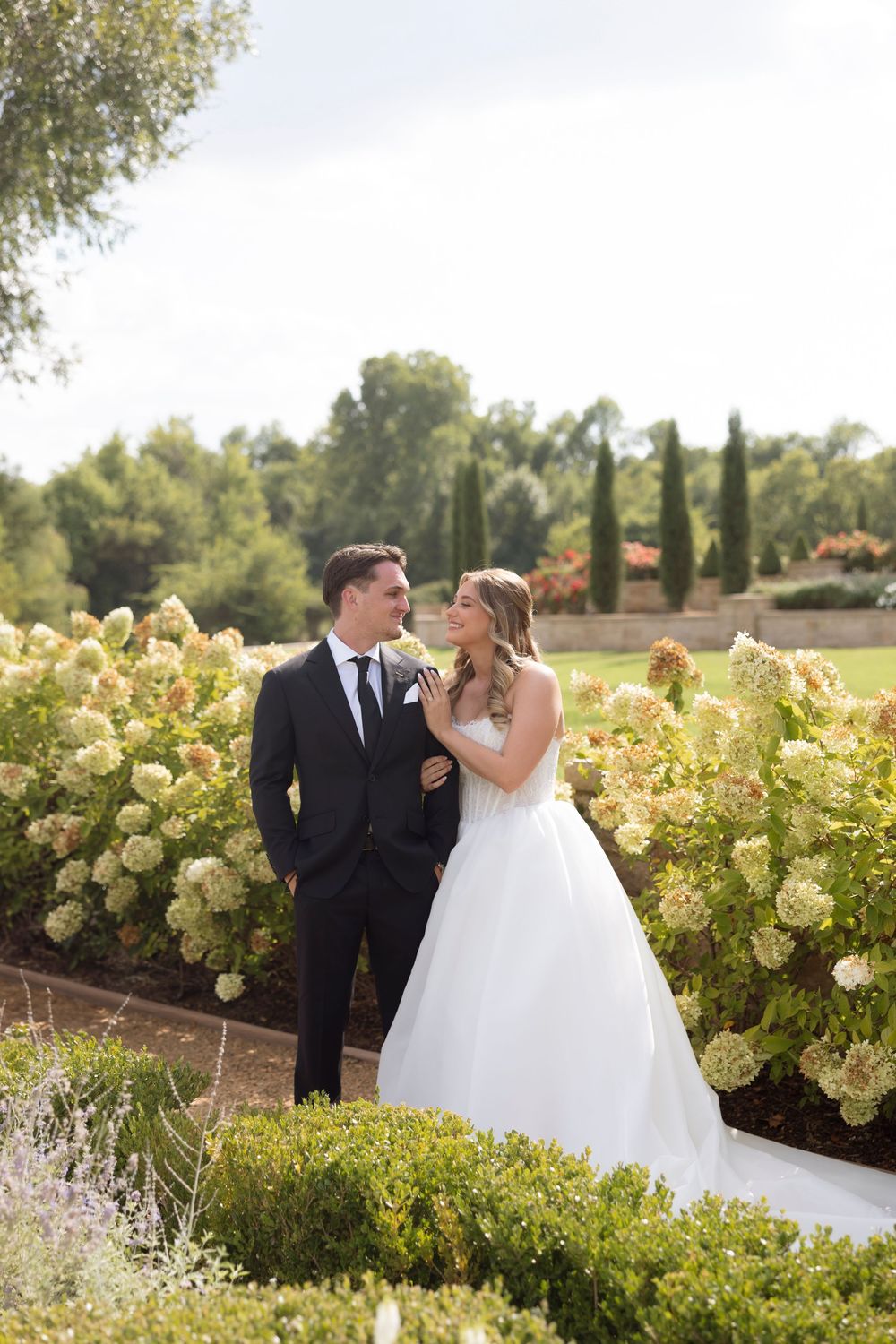A couple in wedding attire shares a romantic moment in a garden setting with blooming hydrangea bushes.
