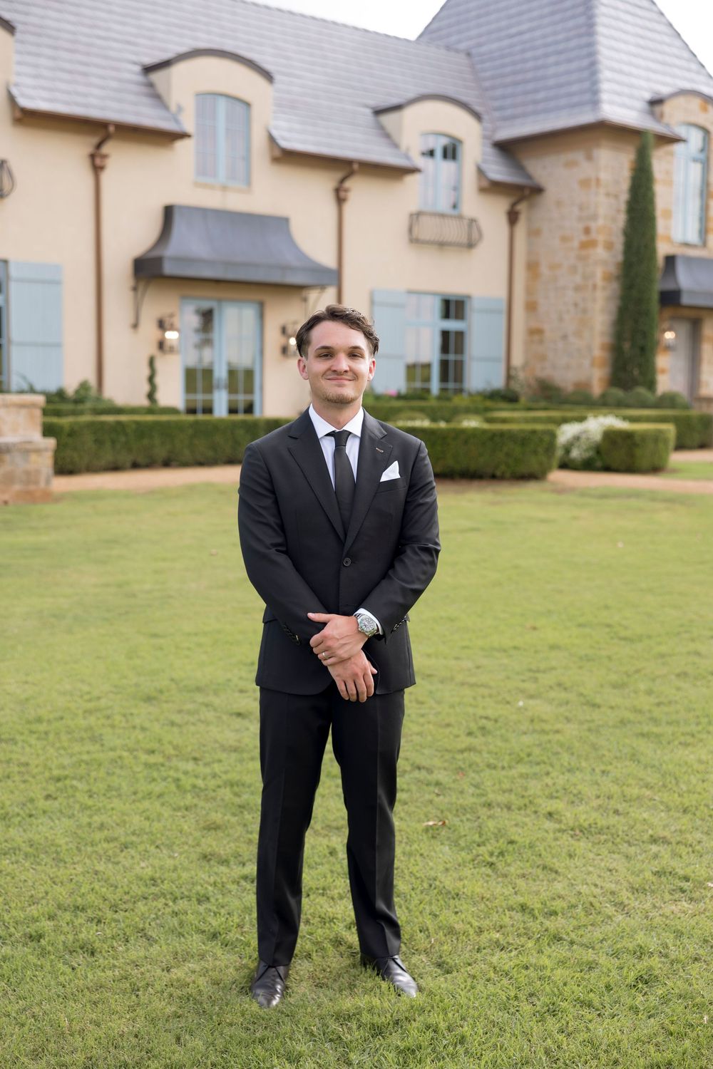 Groom in classic black tuxedo poses on manicured lawn in front of stone mansion.