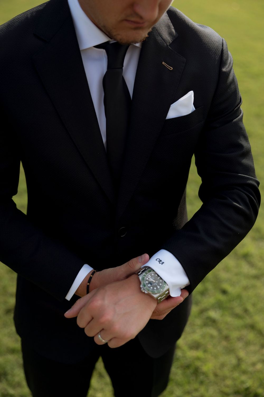 Close-up details of a groom's formal attire including a black suit, watch, and cufflinks against a grassy background.
